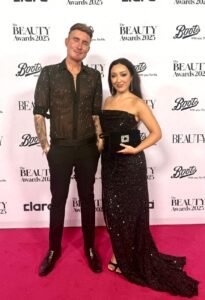 TJ Shirley from TJ tutorials wearing a PHIX Rhinestone shirt and Gucci Pants posing with another guest in front of The Beauty Awards 2025 sponsor backdrop.